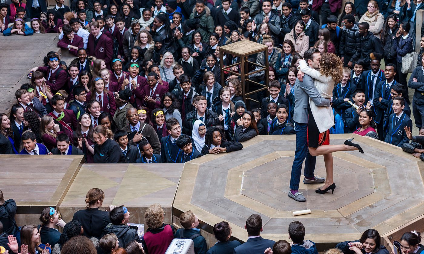 Two actors kiss on stage, on a platform that extends into the audience in the yard