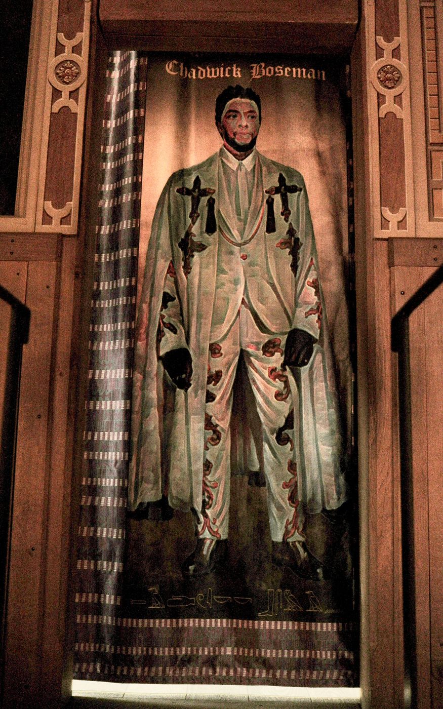 A tapestry of person wearing a white suit and cape decorated with gold crosses and patterns. The tapestry has patterned edges and hangs within a wooden doorframe.