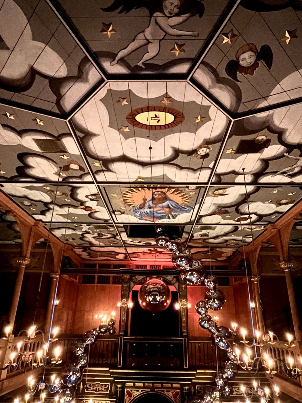The upper gallery and ceiling of a wooden, candlelit theatre. The ceiling is painted grey with white clouds and gold stars. In the centre is an illustration of a celestial figure wearing a long blue dress. A chain of silver balls hangs down from the ceiling and into the gallery.
