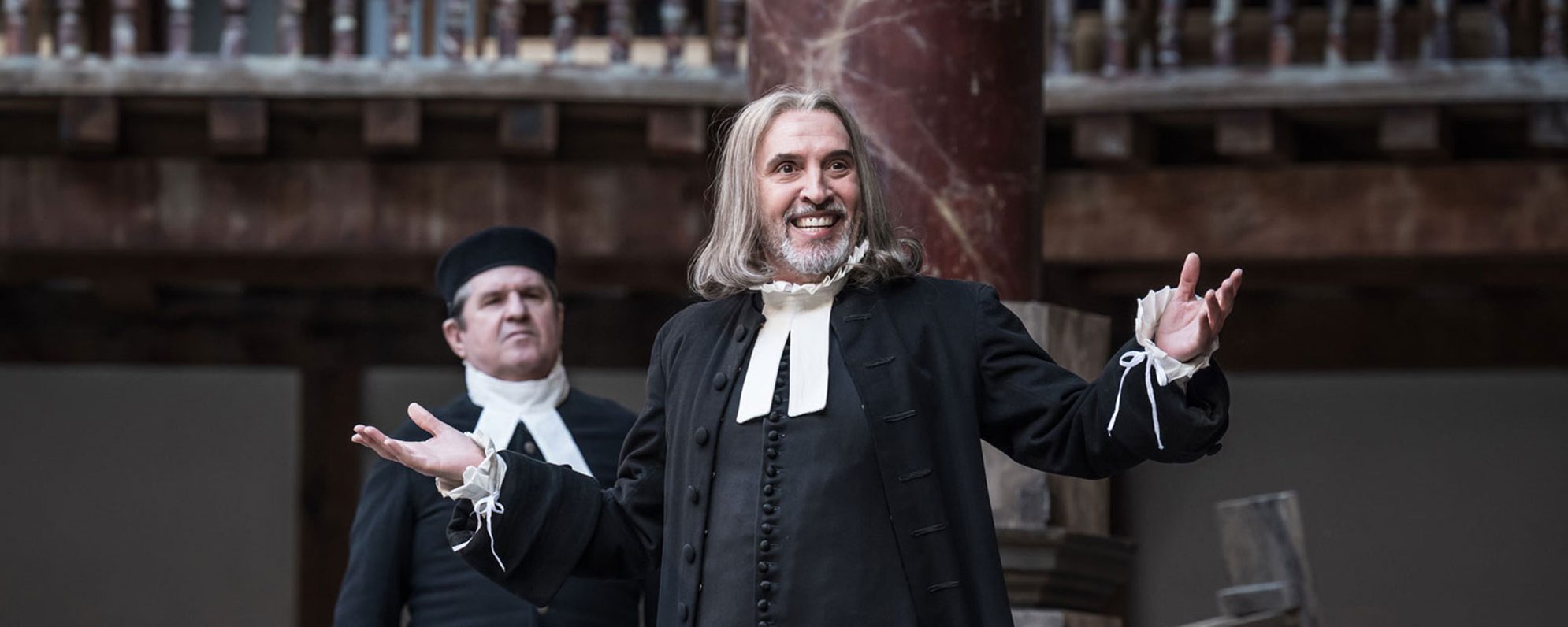 A man on stage at the Globe Theatre, dressed as a Priest, with his arms raised as though he is asking a question.