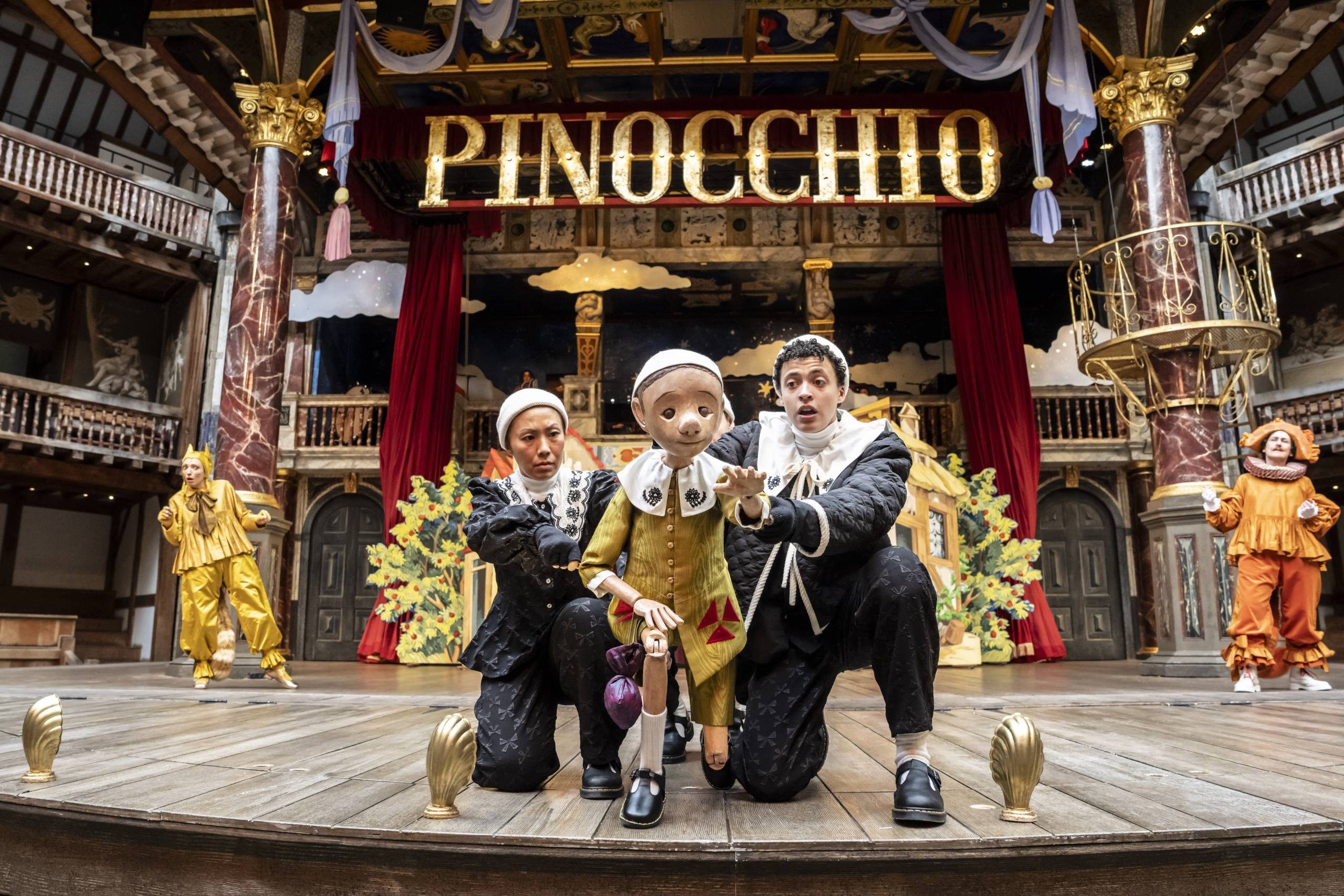 Aya Nakamura as Puppeteer and Lee Braithwaite as Pinocchio hold the wooden puppet of Pinocchio, kneeling centre stage at the Globe Theatre. The puppet of Pinocchio reaches his hand out towards the camera. Above them, gold letters spelling 'Pinocchio' are surrounded by red curtains. Other cast members stand on either side.