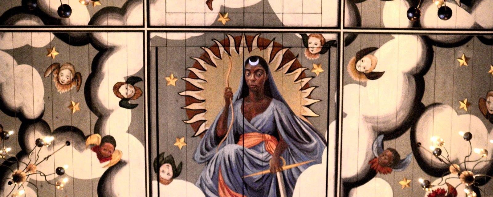 Chandeliers hang from six wooden panels painted grey with white clouds, gold stars and faces with collars and angel wings. In the centre, a person wearing a long, veiled blue dress and a crescent moon on their head sits in front of a circle with pointed edges. The word 'LVNA' is beneath them.