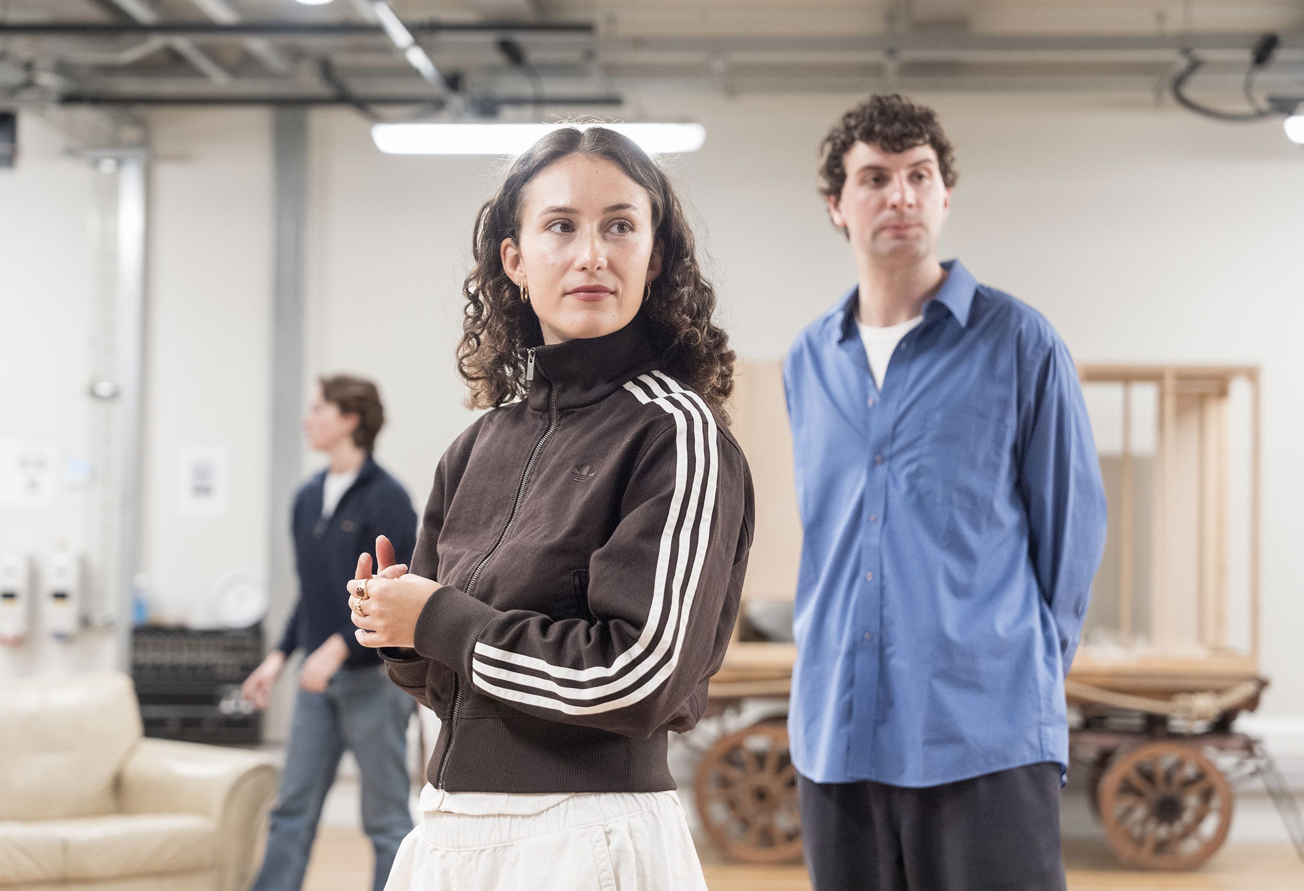 A white, olive-skinned woman with curly brown hair stands and looks across the room. To her right, a white male with curly brown hair stands with his arms behind his back.
