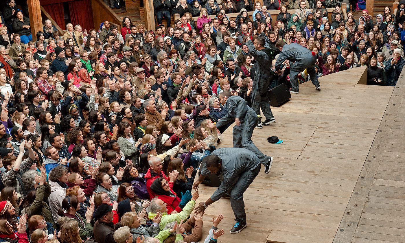 Four actors stand on the edge of a thrust stage, bending down to high five and hold the hands of audience members standing in the Yard in front.