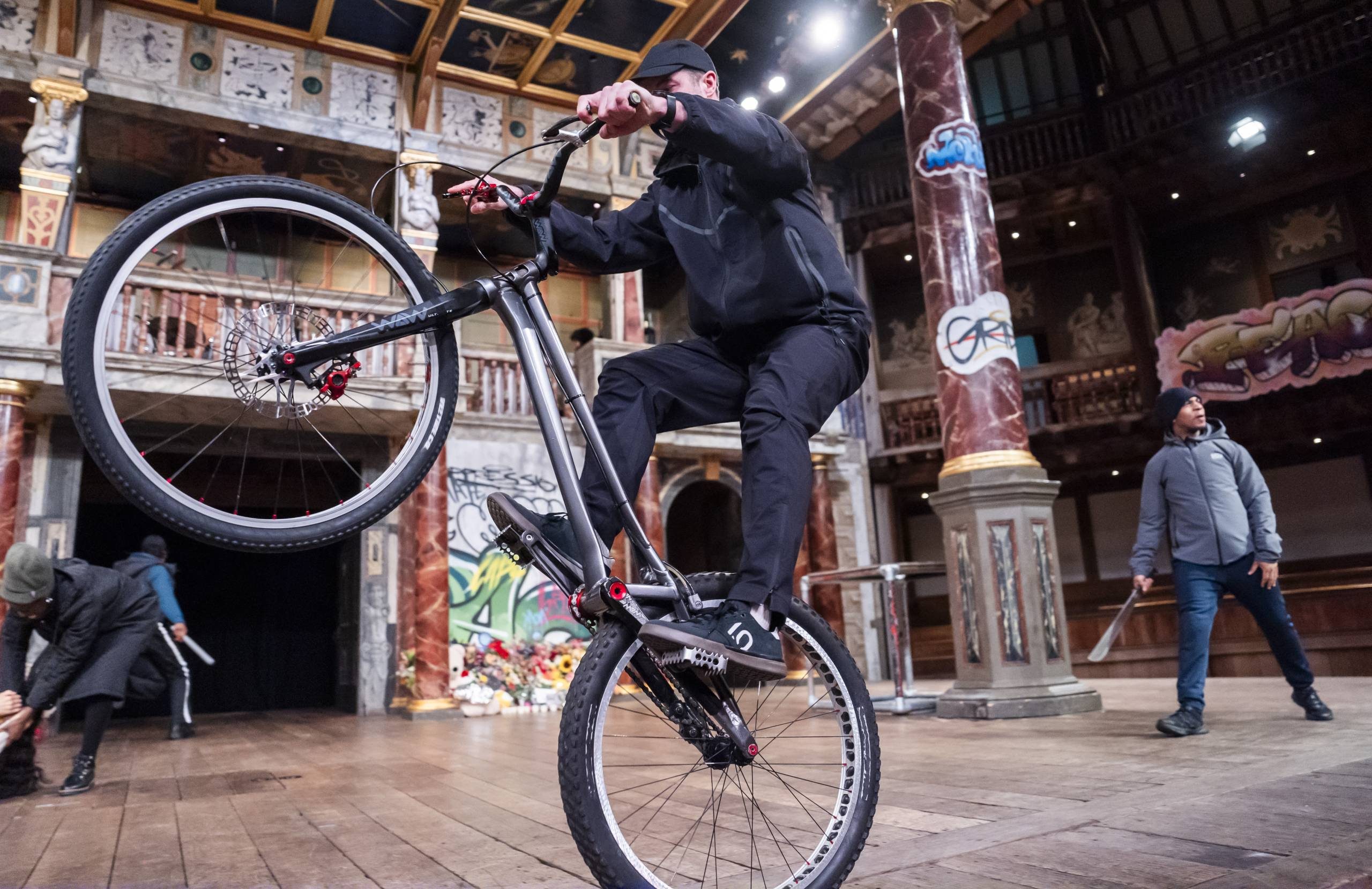 A tall white man with athletic build rides a bike with one wheel in the air on stage. He wears a black tracksuit. There are other cast members, marbled red columns and a balustrade in the background.