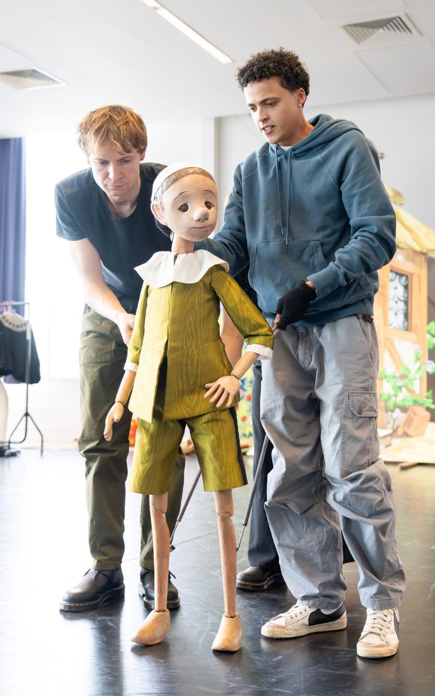 Two actors control a wooden boy puppet. The puppet is wearing a green jacket with a white collar and a white cloth cap.