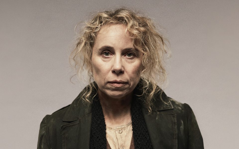 Globe Artistic Director, Michelle Terry, a white woman with long curly hair, looks directly at the camera. She wears a dark dusty trench coat, a wool scarf and a worn cream shirt.