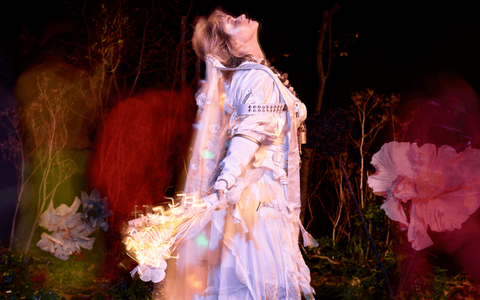A person wearing a white shirt and a pinafore dress with a veil, smiles gently directly into the camera, whilst pointing up with their right hand. They have blonde hair and brown eyes and wear a large bead necklace in various shades of brown. Small orbs of colour appear around them. In the background, various trees are overshadowed by tints of red, yellow and purple.